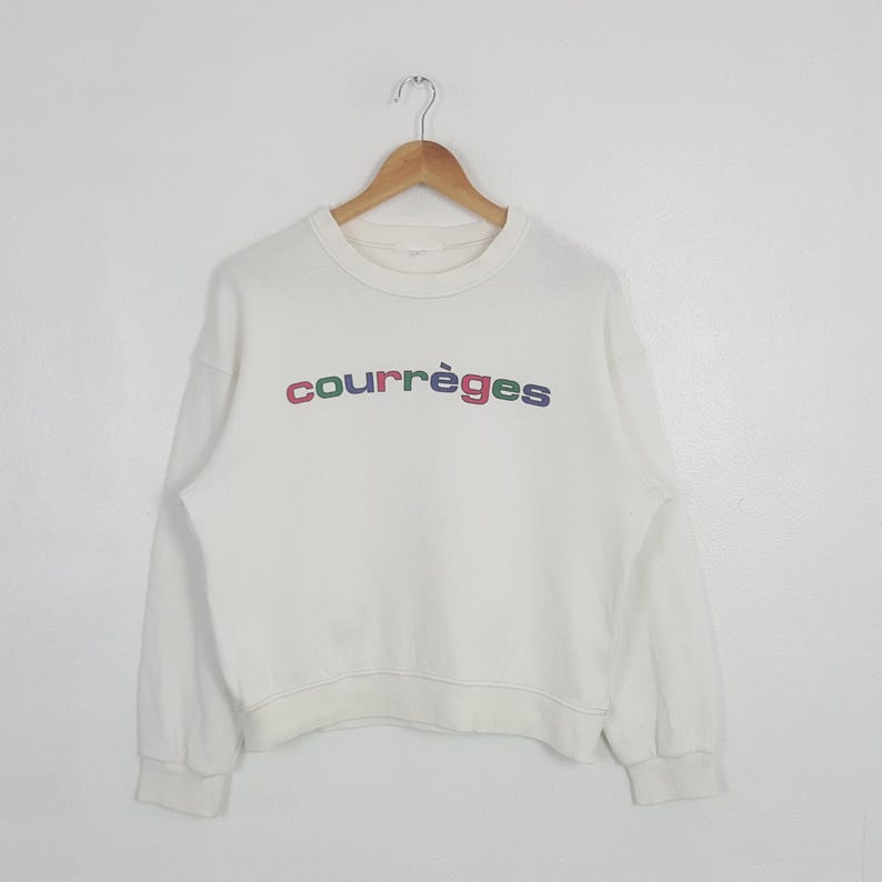 May include: A cream-colored long-sleeve sweatshirt featuring a crew neckline and ribbed cuffs. The word courr&egrave;ges is printed across the chest in lowercase letters, with each letter displayed in a different color including red, green, and blue.