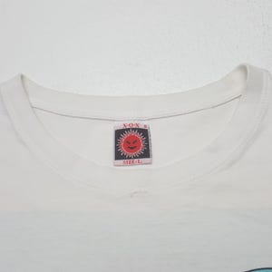 Puede incluir: Camiseta blanca con cuello redondo. La etiqueta presenta el texto "X-O-X" sobre un gr&aacute;fico similar al sol con una cara y el texto "SIZE: L" debajo. La camiseta est&aacute; hecha de un material suave.