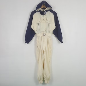 Pode incluir: Macac&atilde;o de esqui vintage creme com mangas e capuz azul marinho. Apresenta cintura com cinto, v&aacute;rios bolsos e fecho de correr frontal. O macac&atilde;o com capuz foi concebido para atividades em climas frios.