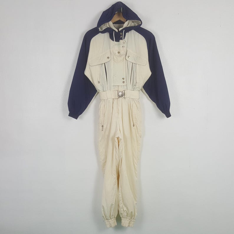 Pode incluir: Macac&atilde;o de esqui vintage creme com mangas e capuz azul marinho. Apresenta cintura com cinto, v&aacute;rios bolsos e fecho de correr frontal. O macac&atilde;o com capuz foi concebido para atividades em climas frios.