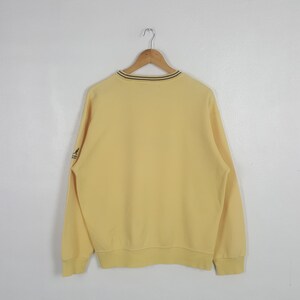 May include: A pale yellow sweatshirt with a crew neck and long sleeves. The neckline has a thin black and brown stripe. A small logo is visible on the left sleeve. The sweatshirt is hanging on a wooden hanger.