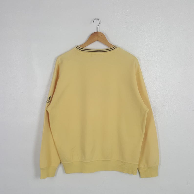 May include: A pale yellow sweatshirt with a crew neck and long sleeves. The neckline has a thin black and brown stripe. A small logo is visible on the left sleeve. The sweatshirt is hanging on a wooden hanger.