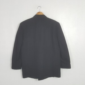 May include: A black blazer hanging on a wooden hanger against a white background. The blazer has a classic design with a notched lapel and two buttons. The sleeves are long, and the fabric appears to be a medium weight.