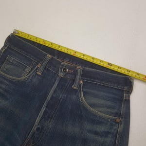 May include: Close-up of blue denim jeans with a button closure and pockets. A yellow measuring tape is laid across the waistband, showing a measurement of approximately 15 inches. The jeans have a classic, worn look.