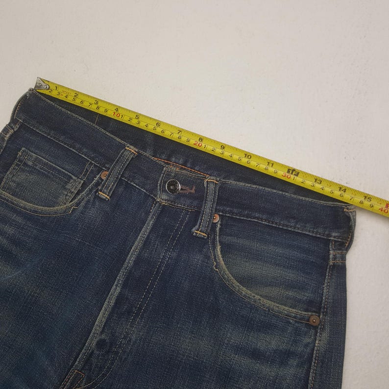 May include: Close-up of blue denim jeans with a button closure and pockets. A yellow measuring tape is laid across the waistband, showing a measurement of approximately 15 inches. The jeans have a classic, worn look.
