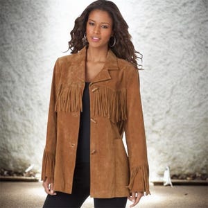 Puede incluir: Chaqueta de ante marrón con flecos en las mangas y el pecho. La chaqueta tiene cuello y botones de madera. La modelo lleva un top y pantalones negros. La chaqueta es de estilo western.