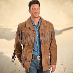 May include: A brown suede jacket with long fringe detailing on the sleeves and chest. The jacket has a button-up front, two front pockets, and a notched collar. The person is wearing a blue denim shirt and blue jeans. A brown leather belt with a silver buckle is visible.