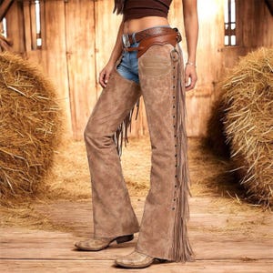 Puede incluir: Chaps de ante marrón con flecos y cinturón a juego. Los chaps presentan un diseño de pierna acampanada y se aseguran con un cinturón. El cinturón tiene un patrón decorativo y una hebilla plateada. Los chaps se usan sobre unos vaqueros azules.