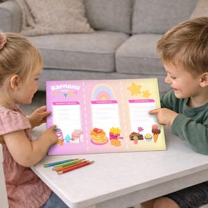 May include: Two children are seated at a white table, looking at a colorful chart. The chart has the text "Barnens" and features illustrations of food and drinks. Colored pencils are on the table. The background includes a couch and decorative items.