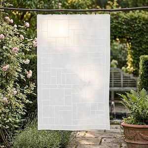 Op de afbeelding: Een rechthoekig wit stoffen paneel met een geometrisch rasterpatroon van elkaar kruisende lijnen. Het semi-transparante materiaal hangt aan een horizontale tak met diverse kleine zwarte clips, waardoor het licht door het textiel valt.