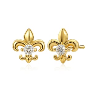 May include: A pair of gold-colored stud earrings. Each earring features a fleur-de-lis design with a sparkling, round, clear gemstone at the center. The earrings are set against a white background.
