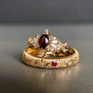 May include: Two gold rings. The top ring features a large, oval, deep red gemstone surrounded by smaller diamonds and white gemstones. The bottom ring is gold with an engraved floral pattern and a small red gemstone.