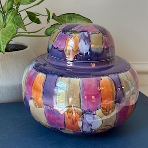 May include: A decorative ceramic jar with a lid. The jar is a deep purple color with vertical rectangular patterns in shades of orange, pink, purple, and tan. The jar is sitting on a blue surface.