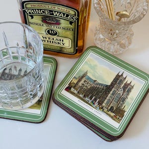 May include: A square coaster featuring a detailed illustration of Westminster Abbey with a green border, placed next to a clear glass tumbler, a bottle of Prince of Wales 10-year-old single vatted malt Welsh whisky, and a crystal glass holder.