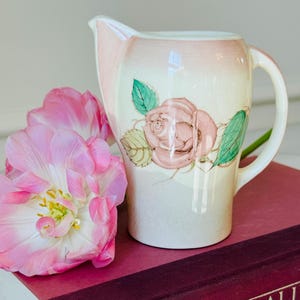May include: A vintage cream-colored pitcher with a pink rose design and a pink rim. The pitcher has a curved handle and a spout. Pink tulips are next to the pitcher, resting on a burgundy book.
