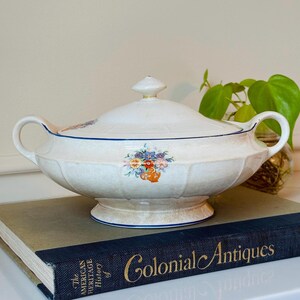 May include: A white ceramic tureen with a lid, featuring a floral design and blue trim. The tureen has handles on either side and sits on a base. The lid has a small knob. The tureen is placed on a book titled "Colonial Antiques".