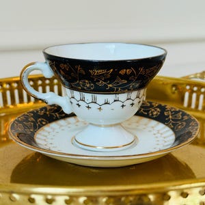 May include: A white porcelain teacup and saucer set featuring a wide black band around the rim and base. The black sections are decorated with intricate gold floral patterns and fine line details. The cup has a pedestal base and a curved handle.