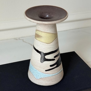May include: A ceramic vase with a wide, dark brown rim and a conical body. The vase features abstract designs in black, yellow, and light blue against a cream-colored background. The vase is approximately 6 inches tall.