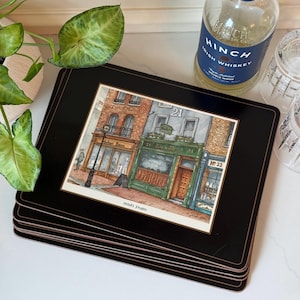 May include: A stack of black rectangular placemats with a watercolor print of Irish shops. The print features buildings with green, brown, and gray facades. A bottle of Hinch Irish Whiskey and two glasses are in the background.