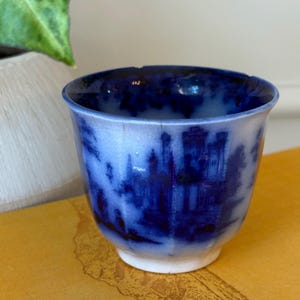 May include: A small, antique blue and white ceramic cup with a landscape design. The cup features a detailed illustration of a castle and buildings. The cup has a white base and a dark blue interior, with visible cracks.