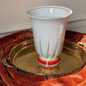 May include: White ceramic vase with a red rim and base, accented with gold lines and a gold tray. The vase has a flared top and a decorative design. The vase is sitting on a gold-colored tray.