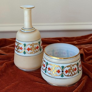 May include: Two-piece set of antique glass bottles. The bottles are beige with a white band featuring gold, red, and green floral designs. Gold bands encircle the top and base of the bottles. The set is displayed on a dark red velvet surface.