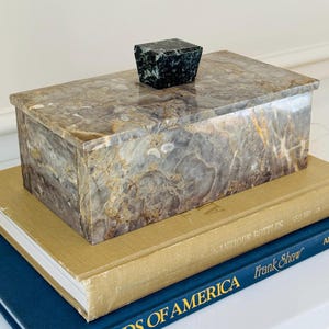 May include: A rectangular stone box with a polished finish featuring swirling patterns of gray, brown, and white. The lid is topped with a small, dark green, trapezoidal stone handle. The box has a smooth, reflective surface and a solid construction.