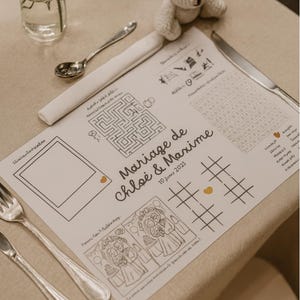 Peut inclure: Un set de table de mariage avec un labyrinthe, un morpion et d'autres jeux. Le set de table porte l'inscription "Mariage de Chloé & Maxime" et la date du "10 Janvier 2023". Un petit ours en peluche crocheté blanc est posé sur la table.