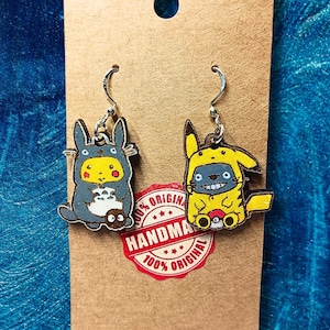 Puede incluir: Pendientes de madera hechos a mano con personajes de dibujos animados. Un pendiente representa una criatura gris y azul, el otro una criatura amarilla. Ambos tienen ganchos plateados. Se muestran en una tarjeta marrón con una pegatina roja "HANDMADE 100% ORIGINAL".
