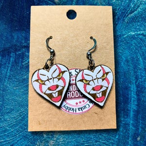 Puede incluir: Pendientes en forma de corazón con un diseño de cara de payaso en rojo, blanco y marrón. Los pendientes cuelgan de ganchos plateados y se presentan en una tarjeta marrón con el texto "Cuska Hobby".
