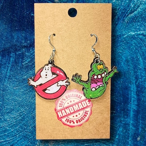 Op de afbeelding: Een paar handgemaakte Ghostbusters-oorbellen. Eén oorbel toont het Ghostbusters-logo in rood en wit. De andere oorbel toont Slimer in groen. De oorbellen zijn bevestigd aan zilverkleurige oorhaakjes. Een sticker leest "100% Handmade 100% Original".