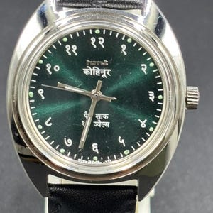 Puede incluir: Primer plano de un reloj de pulsera con caja plateada y esfera verde oscuro. El reloj tiene números y agujas blancos, con texto en idioma extranjero. Correa de cuero negro.