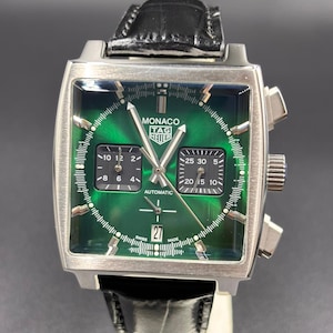 K&ouml;nnte beinhalten: Eine quadratische TAG Heuer Monaco Automatikuhr mit gr&uuml;nem Sonnenschliff-Zifferblatt, zwei schwarzen quadratischen Hilfszifferbl&auml;ttern und einer Datumsanzeige auf der Sechs-Uhr-Position. Die Uhr hat silberfarbene Zeiger und Indizes sowie ein schwarzes Lederarmband.