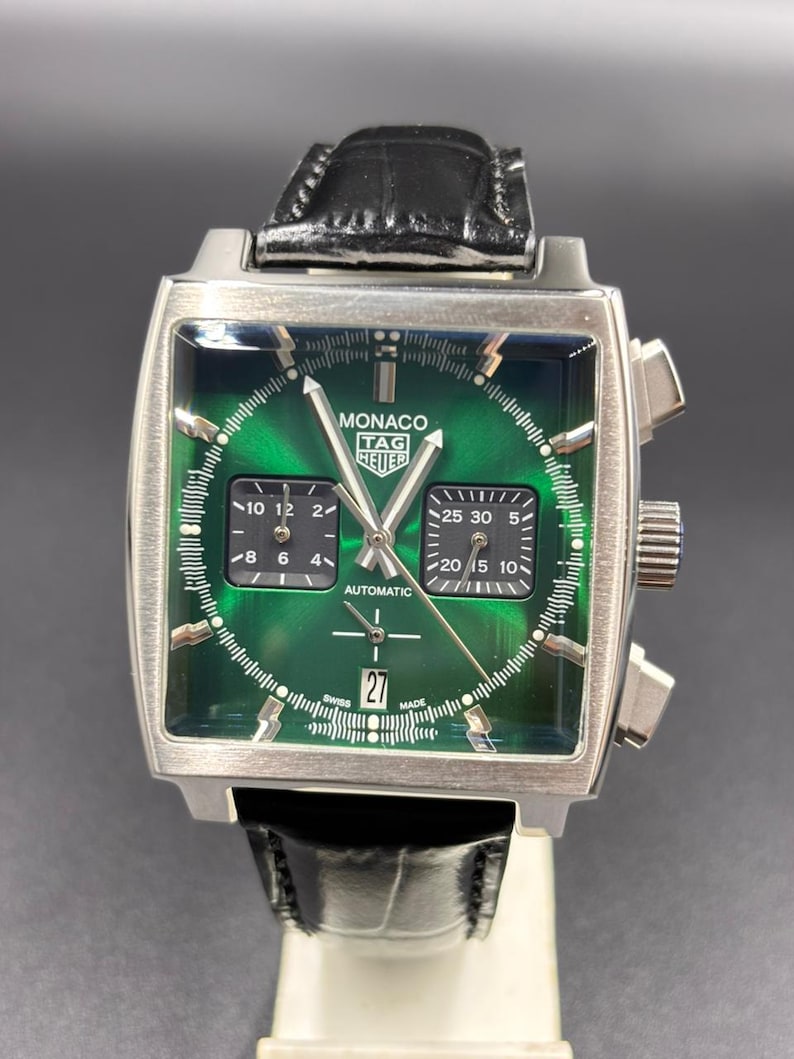 K&ouml;nnte beinhalten: Eine quadratische TAG Heuer Monaco Automatikuhr mit gr&uuml;nem Sonnenschliff-Zifferblatt, zwei schwarzen quadratischen Hilfszifferbl&auml;ttern und einer Datumsanzeige auf der Sechs-Uhr-Position. Die Uhr hat silberfarbene Zeiger und Indizes sowie ein schwarzes Lederarmband.