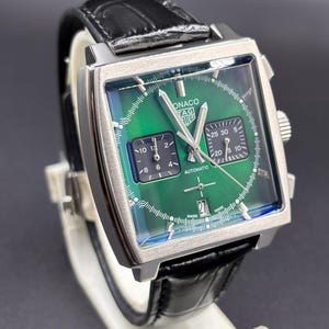 K&ouml;nnte beinhalten: Eine quadratische TAG Heuer Monaco Armbanduhr mit gr&uuml;nem Sonnenschliff-Zifferblatt, zwei schwarzen quadratischen Hilfszifferbl&auml;ttern, silberfarbenen Zeigern und einer Datumsanzeige auf der Sechs-Uhr-Position. Sie hat ein schwarzes Lederarmband.