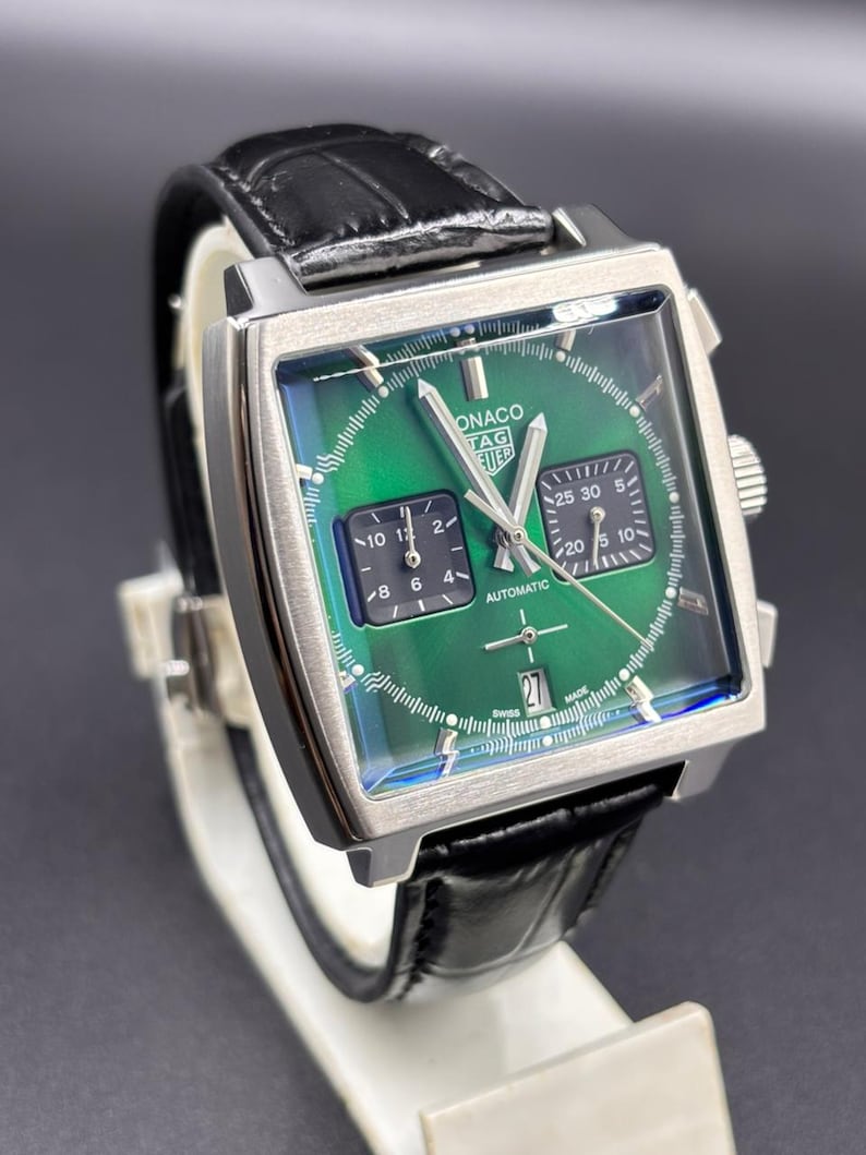 K&ouml;nnte beinhalten: Eine quadratische TAG Heuer Monaco Armbanduhr mit gr&uuml;nem Sonnenschliff-Zifferblatt, zwei schwarzen quadratischen Hilfszifferbl&auml;ttern, silberfarbenen Zeigern und einer Datumsanzeige auf der Sechs-Uhr-Position. Sie hat ein schwarzes Lederarmband.