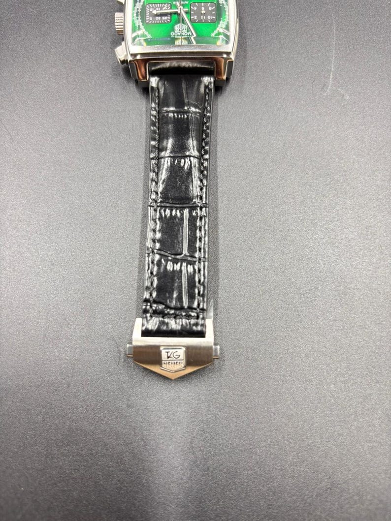 K&ouml;nnte beinhalten: Eine Armbanduhr mit quadratischem Geh&auml;use und leuchtend gr&uuml;nem Zifferblatt, das zwei schwarze Hilfszifferbl&auml;tter und wei&szlig;e Markierungen aufweist. Die Uhr hat ein poliertes silberfarbenes Metallgeh&auml;use und ein schwarzes strukturiertes Lederarmband mit einer silberfarbenen Metallschlie&szlig;e, die das TAG Heuer-Logo zeigt.