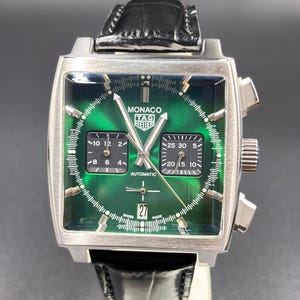 K&ouml;nnte beinhalten: Eine quadratische TAG Heuer Monaco Armbanduhr mit gr&uuml;nem Sonnenschliff-Zifferblatt, zwei schwarzen quadratischen Hilfszifferbl&auml;ttern und einer Datumsanzeige auf der Sechs-Uhr-Position. Die Uhr hat silberfarbene Zeiger, Indizes, ein schwarzes Lederarmband und ein Edelstahlgeh&auml;use.