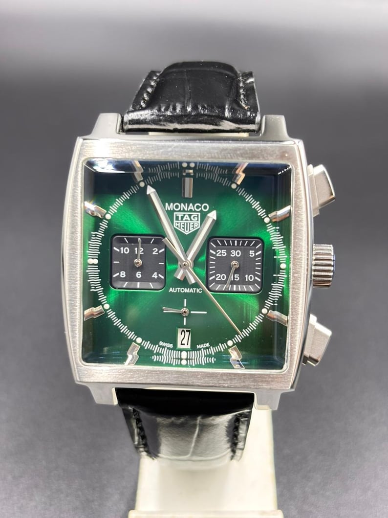 K&ouml;nnte beinhalten: Eine quadratische TAG Heuer Monaco Armbanduhr mit gr&uuml;nem Sonnenschliff-Zifferblatt, zwei schwarzen quadratischen Hilfszifferbl&auml;ttern und einer Datumsanzeige auf der Sechs-Uhr-Position. Die Uhr hat silberfarbene Zeiger, Indizes, ein schwarzes Lederarmband und ein Edelstahlgeh&auml;use.