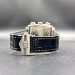 K&ouml;nnte beinhalten: Ein schwarzes Lederuhrenarmband mit einer silberfarbenen Metall-Faltschlie&szlig;e, die das TAG Heuer-Logo tr&auml;gt. Das Armband hat eine strukturierte Oberfl&auml;che und sichtbare N&auml;hte und ist an der R&uuml;ckseite einer quadratischen Uhr mit silbernen Dr&uuml;ckern befestigt.