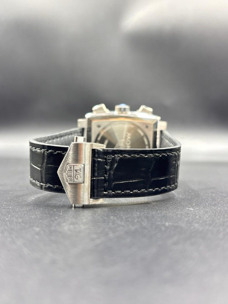 K&ouml;nnte beinhalten: Ein schwarzes Lederuhrenarmband mit einer silberfarbenen Metall-Faltschlie&szlig;e, die das TAG Heuer-Logo tr&auml;gt. Das Armband hat eine strukturierte Oberfl&auml;che und sichtbare N&auml;hte und ist an der R&uuml;ckseite einer quadratischen Uhr mit silbernen Dr&uuml;ckern befestigt.
