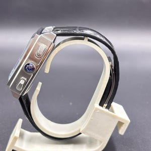 K&ouml;nnte beinhalten: Eine rechteckige Armbanduhr mit einem silberfarbenen Metallgeh&auml;use und einem schwarzen, strukturierten Lederarmband. Die Seite des Geh&auml;uses zeigt zwei rechteckige Dr&uuml;cker und eine zentrale Krone mit einem blauen Emblem. Seitenansicht.