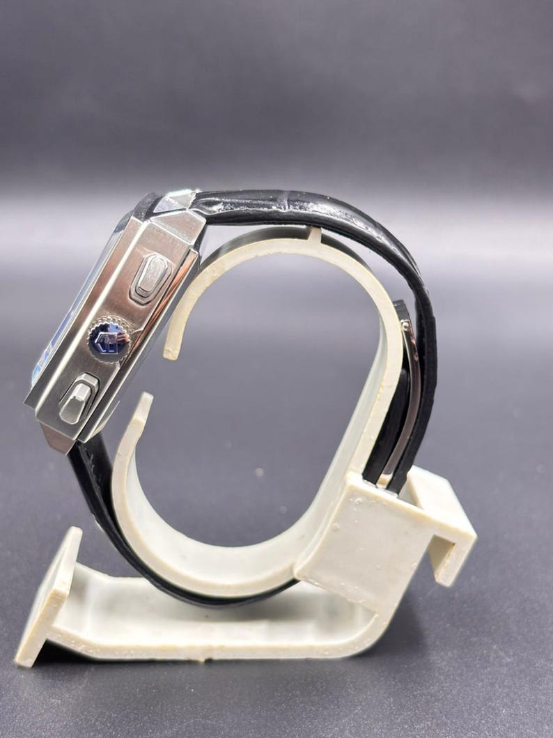 K&ouml;nnte beinhalten: Eine rechteckige Armbanduhr mit einem silberfarbenen Metallgeh&auml;use und einem schwarzen, strukturierten Lederarmband. Die Seite des Geh&auml;uses zeigt zwei rechteckige Dr&uuml;cker und eine zentrale Krone mit einem blauen Emblem. Seitenansicht.