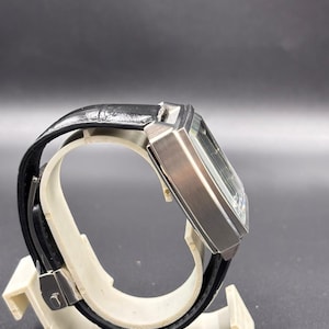 K&ouml;nnte beinhalten: Eine rechteckige Armbanduhr mit einem Geh&auml;use aus geb&uuml;rstetem silberfarbenem Metall und einem schwarzen Lederarmband. Die Uhr zeigt eine Seitenansicht mit dem dicken, kantigen Geh&auml;use und dem strukturierten schwarzen Band mit Metallschlie&szlig;e.