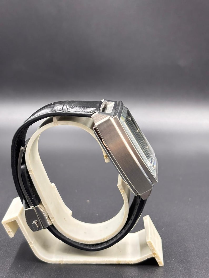 K&ouml;nnte beinhalten: Eine rechteckige Armbanduhr mit einem Geh&auml;use aus geb&uuml;rstetem silberfarbenem Metall und einem schwarzen Lederarmband. Die Uhr zeigt eine Seitenansicht mit dem dicken, kantigen Geh&auml;use und dem strukturierten schwarzen Band mit Metallschlie&szlig;e.