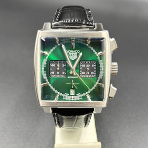 K&ouml;nnte beinhalten: Eine quadratische TAG Heuer Monaco Armbanduhr mit gr&uuml;nem Sonnenschliff-Zifferblatt, zwei schwarzen quadratischen Hilfszifferbl&auml;ttern und einer Datumsanzeige auf der Sechs-Uhr-Position. Sie hat silberfarbene Zeiger, ein schwarzes Lederarmband und den Schriftzug Monaco TAG Heuer.