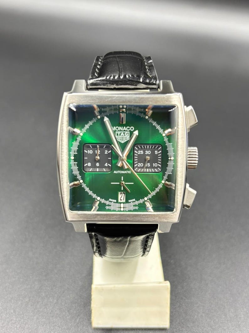 K&ouml;nnte beinhalten: Eine quadratische TAG Heuer Monaco Armbanduhr mit gr&uuml;nem Sonnenschliff-Zifferblatt, zwei schwarzen quadratischen Hilfszifferbl&auml;ttern und einer Datumsanzeige auf der Sechs-Uhr-Position. Sie hat silberfarbene Zeiger, ein schwarzes Lederarmband und den Schriftzug Monaco TAG Heuer.