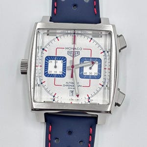 Może przedstawiać: Srebrny zegarek chronograf z kwadratową kopertą, białą tarczą, dwiema niebieskimi subtarczami i czerwonymi akcentami. Na tarczy widnieje napis Monaco Heuer Automatic Chronograph Swiss Made. Niebieski perforowany sk&oacute;rzany pasek z czerwonymi przeszyciami.