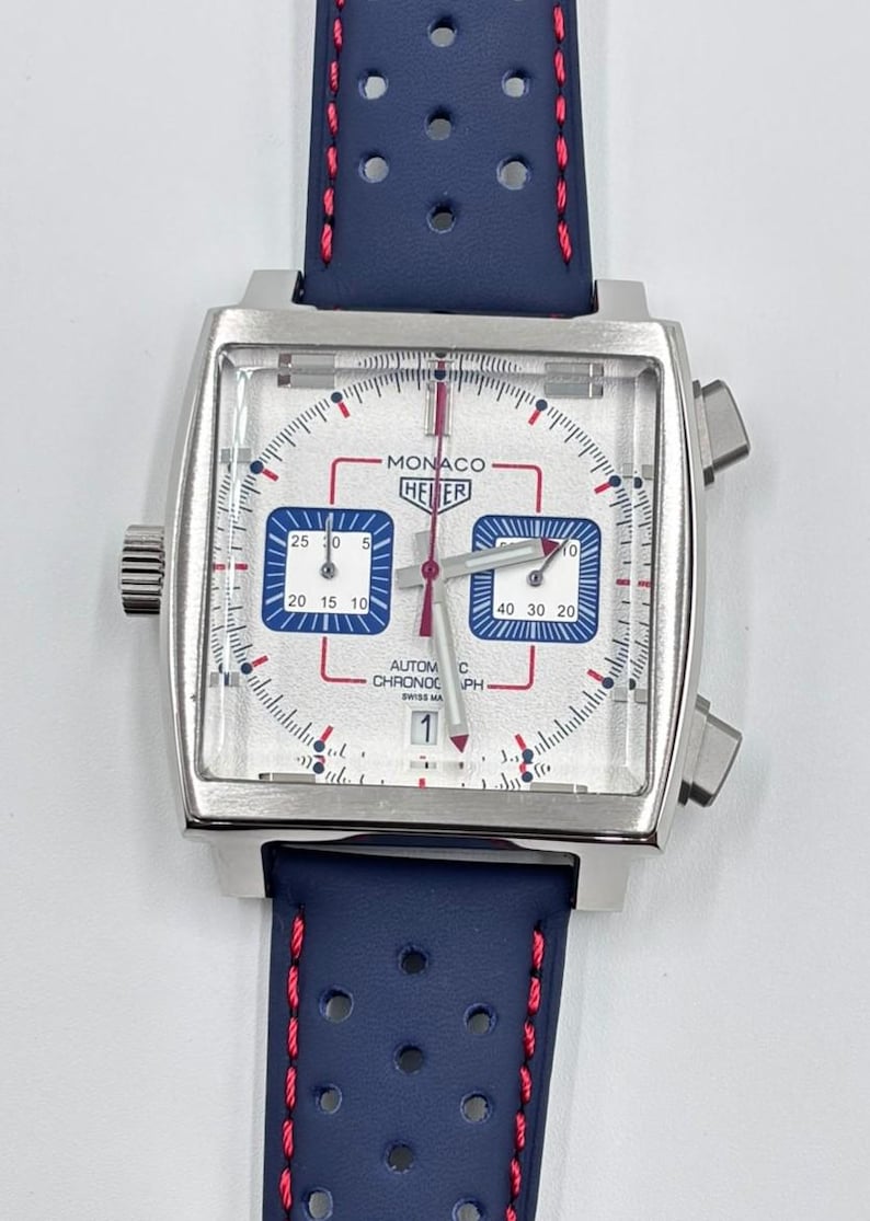 Może przedstawiać: Srebrny zegarek chronograf z kwadratową kopertą, białą tarczą, dwiema niebieskimi subtarczami i czerwonymi akcentami. Na tarczy widnieje napis Monaco Heuer Automatic Chronograph Swiss Made. Niebieski perforowany sk&oacute;rzany pasek z czerwonymi przeszyciami.