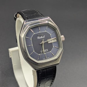 Puede incluir: Un reloj de pulsera vintage plateado con esfera azul oscuro y correa de cuero negro. La esfera muestra el día y la fecha, con la marca "Kailash" visible. El reloj se exhibe sobre un soporte blanco.